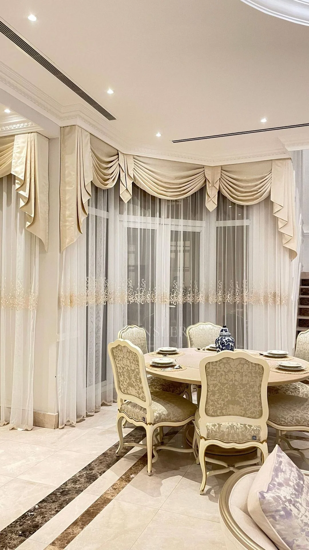 Dining table and tall ivory curtains with ornamental upper folds.